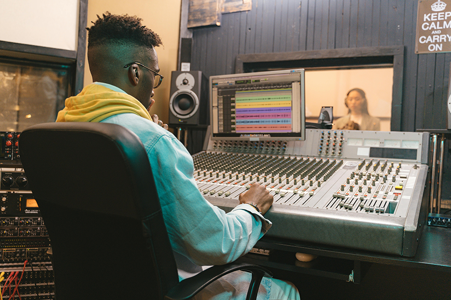 A music producer sitting at a large mixing console in a recording studio, focused on adjusting controls while a vocalist stands in a sound booth in the background, illustrating the process of music production and recording.